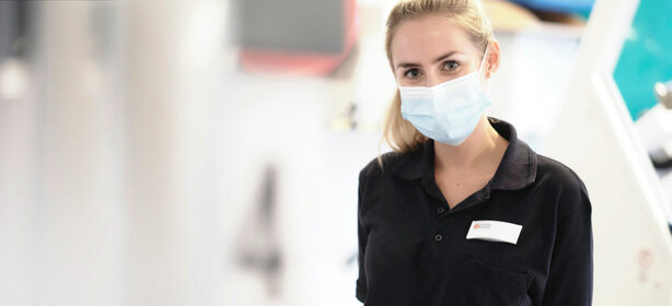 Junge Frau mit blonden Haaren, medizinischer Gesichtsmaske und Poloshirt mit Namensschild im hellen Innenraum