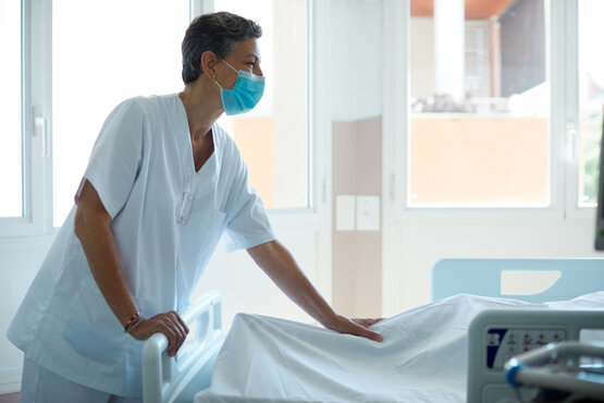 Pflegekraft mit kurzen Haaren und blauer Maske steht in weißer Arbeitskleidung vor Krankenbett im hellen Zimmer, Tageslicht durch große Fenster