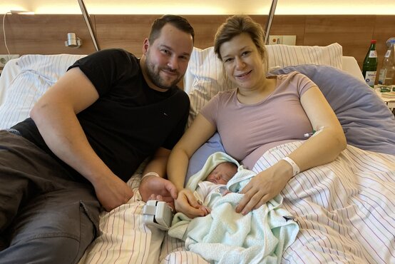 Junges Elternpaar mit Neugeborenem im Krankenhausbett, Mutter hält Baby, Vater hält ihre Hand, Familienglück nach Geburt