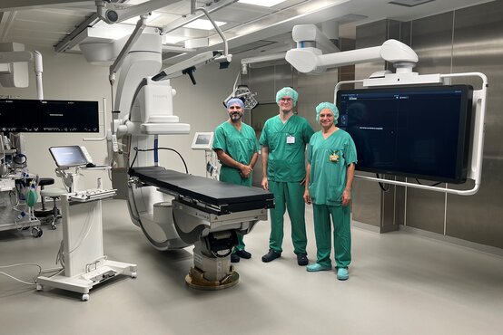 Drei Personen in grüner OP-Kleidung im modernen Hybrid-Operationssaal mit bildgebender Medizintechnik und Monitoren