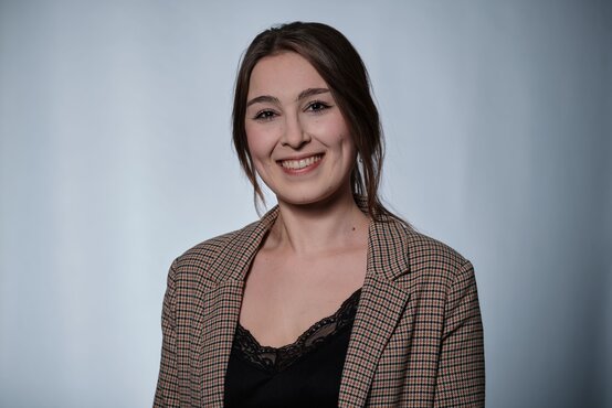 Junge Frau mit langen braunen Haaren im karierten Blazer, lächelt freundlich vor neutral-grauem Hintergrund, Porträtfoto