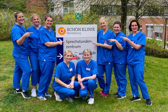 Acht Frauen in blauer medizinischer Kleidung vor Schild der Schön Klinik Rendsburg, Teamfoto auf grünem Rasen