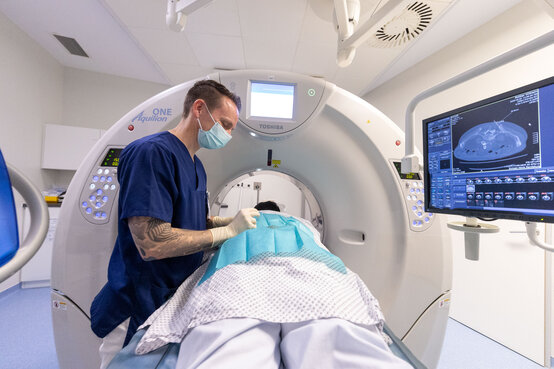 Mann in blauem Kittel mit Mundschutz steht neben Patientin auf Liege, die in CT-Gerät geschoben wird, Monitor mit Querschnittsbild rechts