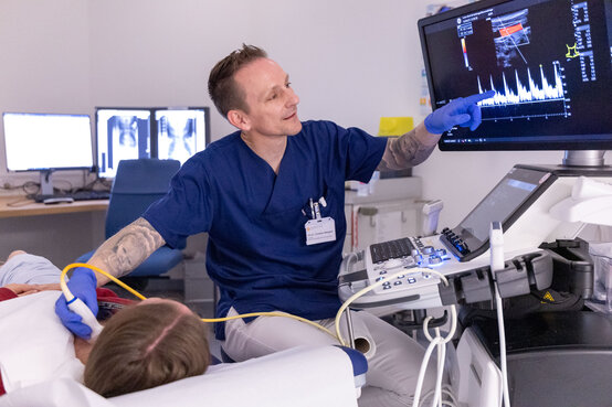 Mann in blauer medizinischer Kleidung führt Ultraschalluntersuchung am Hals einer liegenden Person durch, zeigt auf Monitor mit Ultraschallbild