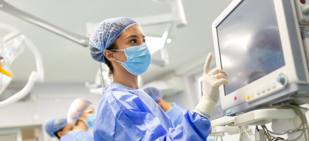 Person in blauer OP-Kleidung und Maske berührt mit Handschuh einen Monitor im hellen Operationssaal, weitere Personen und Geräte im Hintergrund
