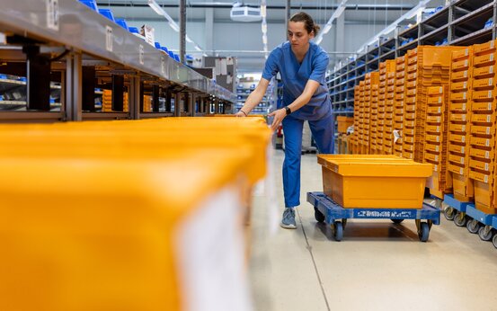 Mitarbeiter in blauer Arbeitskleidung schiebt Plattform mit orangefarbenen Kisten durch Apothekenlager