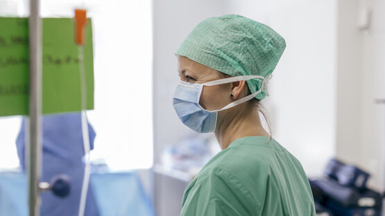 Pflegekraft in grüner OP-Kleidung, Haube und Maske steht im hellen OP-Raum, Infusionsstange und medizinische Geräte im Hintergrund