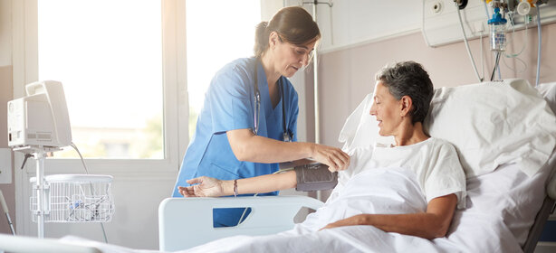 Pflegekraft in blauer Kleidung legt Patientin im weißen T-Shirt im Krankenbett eine Blutdruckmanschette am linken Oberarm an