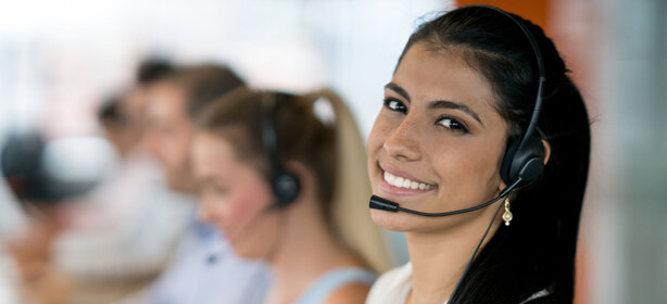 Freundliche Frau mit Headset, lächelt in die Kamera, im Hintergrund weitere Mitarbeitende