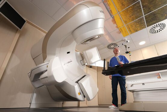 Person in blauer Kleidung mit Klemmbrett steht neben großem medizinischem-Gerät in hellem Raum, Liege mit medizinischem Zubehör davor