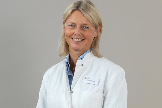 Dr. Simone Pfeuffer mit schulterlangem blondem Haar im weißen Arztkittel mit Namensschild vor grauem Hintergrund