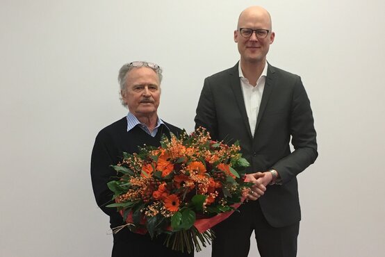 Mann hält großen Blumenstrauß, jüngerer Mann im Anzug steht daneben, beide vor weißer Wand