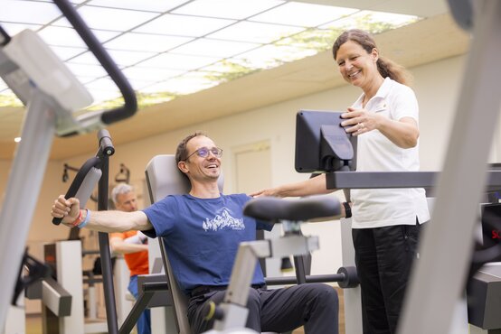 Mann trainiert an Brustpresse, Physiotherapeutin im weißen Polohemd unterstützt ihn