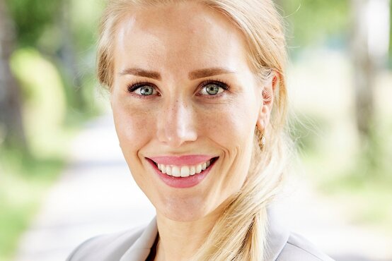 Porträt von Dr. Sophie Giessner, junge Frau mit blondem Haar im hellen Blazer, draußen vor grünem Hintergrund
