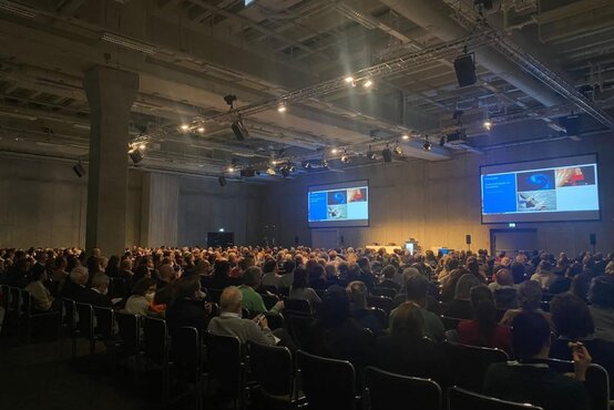 Großer Konferenzsaal mit vielen Teilnehmenden bei medizinischem Fachkongress, Präsentationsfolien auf Leinwänden sichtbar