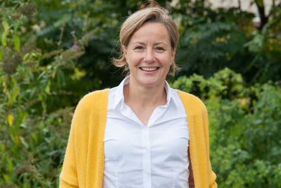 Lächelnde Frau mit kurzem, hellbraunem Haar in weißer Bluse und gelber Strickjacke vor grünem, unscharfem Hintergrund