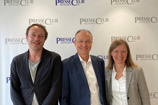 Drei Personen vor Fotowand mit Schriftzug „PresseClub München e.V.“, lächeln in die Kamera, formelle Kleidung