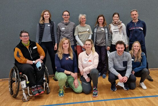 Gruppenfoto von elf Erwachsenen in Freizeitkleidung, eine Person im Rollstuhl, alle posieren in Sporthalle vor grauer Wand