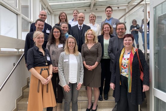 Gruppenfoto von 15 Personen auf Treppe in hellem, verglasten Innenbereich, alle lächeln in die Kamera