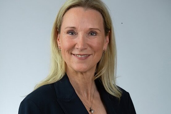 Porträt einer lächelnden Frau mit blondem Haar im dunklen Blazer und weißer Bluse vor hellem Hintergrund