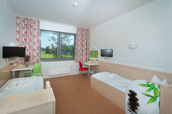 Helles, modernes Klinik-Doppelzimmer mit zwei Betten, großen Fenstern, bunten Vorhängen und Schreibtischen für Patienten