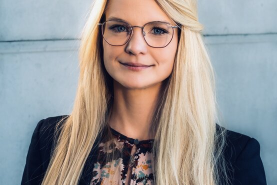 Junge Frau mit langem, blondem Haar und Brille, schwarze Jacke, gemusterte Bluse, vor hellgrauer Betonwand, frontal blickend
