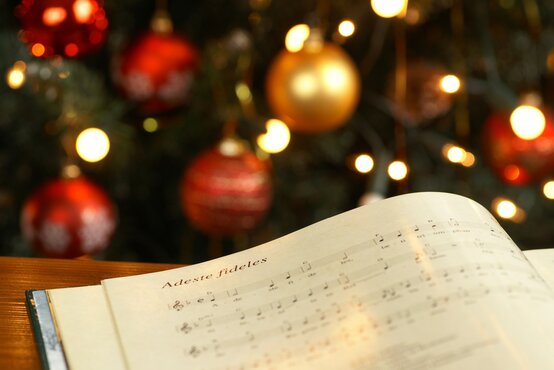 Geöffnetes Notenbuch mit „Adeste fideles“ auf Holztisch, im Hintergrund festlich geschmückter Weihnachtsbaum mit Lichterkette