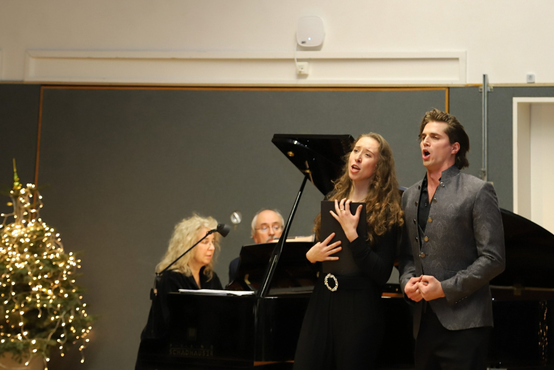 Zwei singende Personen vor schwarzem Flügel, im Hintergrund zwei Menschen am Klavier, links leuchtender Weihnachtsbaum