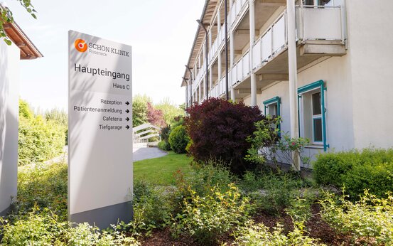 Großes Schild „Schön Klinik Roseneck Haupteingang Haus C“ vor mehrstöckigem Gebäude mit Balkonen und Grünpflanzen