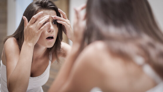 Junge Frau mit langen braunen Haaren betrachtet ihr Gesicht besorgt im Spiegel und berührt ihre Stirn mit beiden Händen
