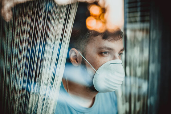 Junger Mann mit Gesichtsmaske steht nachdenklich hinter Fenster mit Vorhang, warmes Licht
