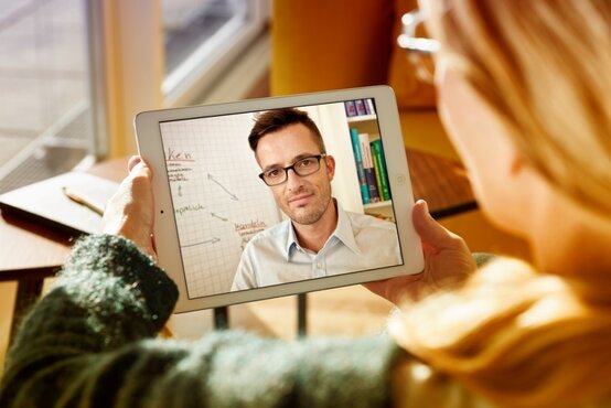 Person hält Tablet mit laufendem Videoanruf, zweite Person auf Bildschirm sichtbar, im Hintergrund Whiteboard und Bücherregale