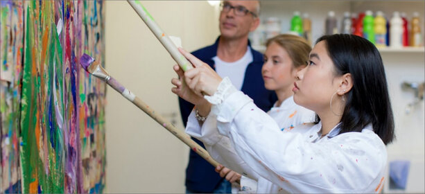 Zwei junge Menschen malen im weißen Kittel mit Pinseln an bunter Leinwand, Therapeut beobachtet, Kunsttherapie-Raum