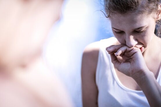 Junge Frau mit braunen Haaren in weißem Top steht nachdenklich und besorgt in hellem Raum, stützt Kopf auf die Hand