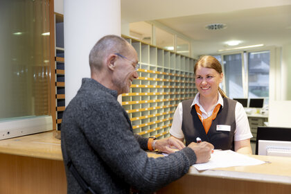 Freundliche Mitarbeiterin am Empfang der Klinik begrüßt älteren Patienten, moderner und heller Empfangsbereich