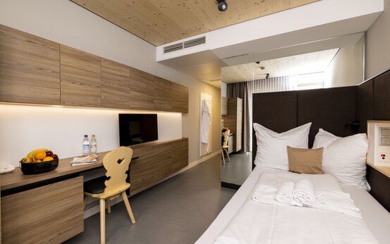 Helles Komfortzimmer mit Einzelbett, Schreibtisch, Fernseher, Obstschale, Wasserflaschen und Holzdecke in der Schön Klinik Roseneck