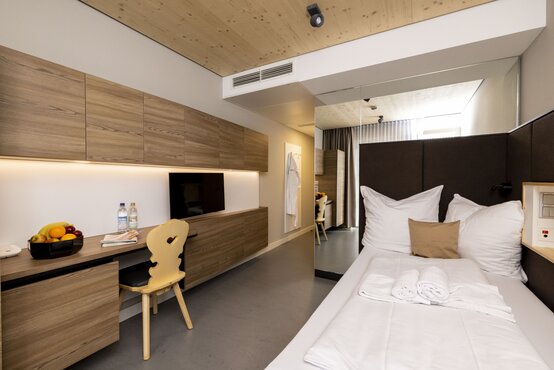Helles Komfortzimmer mit Einzelbett, Schreibtisch, Fernseher, Obstschale, Wasserflaschen und Holzdecke in der Schön Klinik Roseneck