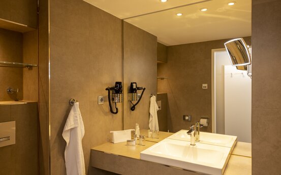 Modernes Bad der Schön Klinik Roseneck mit weißem Waschbecken, großem Spiegel, Handtuch, Fön und warmem Licht