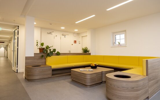 Heller Lounge-Bereich mit L-förmiger Sitzbank mit gelben Polstern, zwei Holztischen, Pflanzen und Obstschale in der Schön Klinik Roseneck
