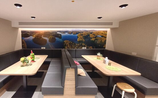 Moderner Aufenthaltsraum mit zwei großen Holztischen, dunklen Polsterbänken, Pflanzen, Wandbild mit Seenlandschaft und Hocker