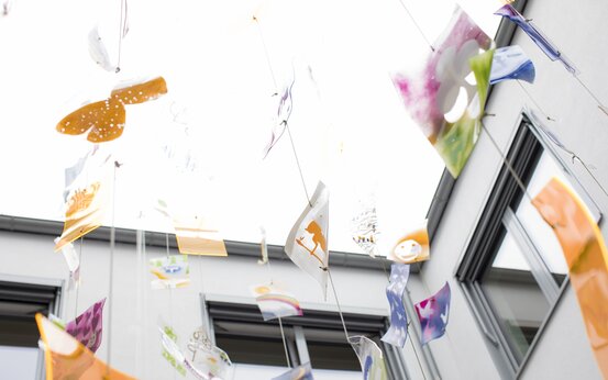 Bunte, bemalte Kunststoffplatten hängen an Schnüren zwischen Gebäudefenstern im Freien vor hellem Himmel