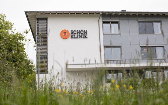 Moderne Fassade der Schön Klinik Roseneck mit weißer und grauer Außenwand, orangem Klinik-Logo, Fenstern, Gräsern und gelben Blumen