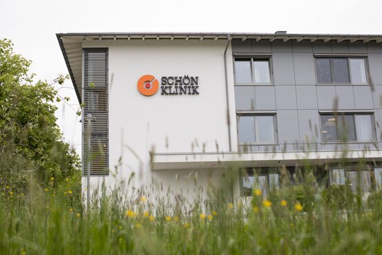 Moderne Fassade der Schön Klinik Roseneck mit weißer und grauer Außenwand, orangem Klinik-Logo, Fenstern, Gräsern und gelben Blumen