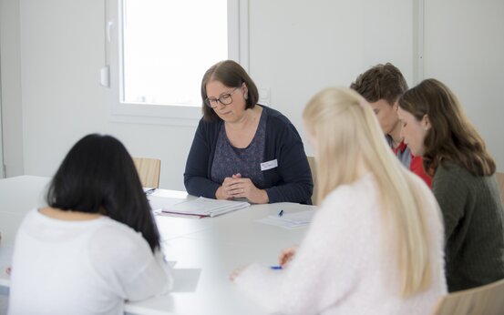 Lehrerin mit Brille leitet kleine Lerngruppe aus vier Jugendlichen an weißem Tisch in hellem Raum, Lernmaterialien und Fenster im Hintergrund