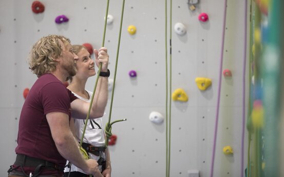 Zwei Personen mit Klettergurten sichern sich in einer Kletterhalle, bunte Kletterwand im Hintergrund