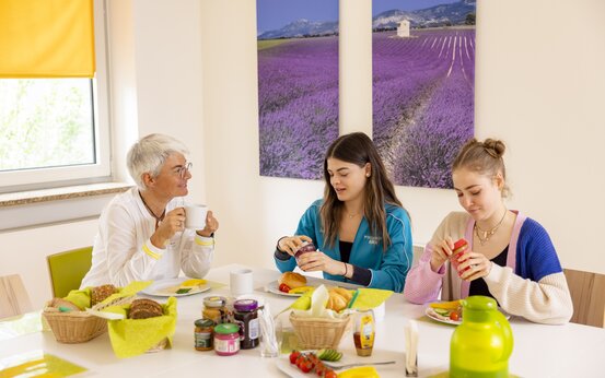 Drei Frauen unterschiedlichen Alters frühstücken gemeinsam an einem hellen Esstisch mit Brötchen, Aufstrichen und frischem Gemüse