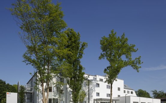 Modernes Klinikgebäude Haus Rosenheim mit weißer Fassade, Zugang, Grünflächen und Bäumen