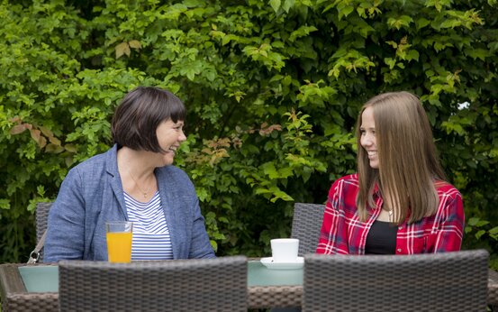 Zwei Frauen sitzen lachend an Gartentisch aus Rattanmöbeln im Grünen, Glas Orangensaft und Kaffeetasse vor sich
