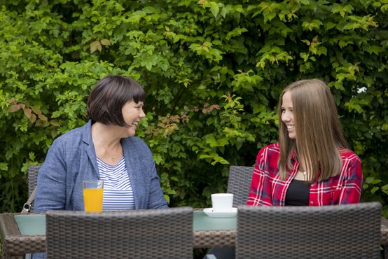 Zwei Frauen sitzen lachend an Gartentisch aus Rattanmöbeln im Grünen, Glas Orangensaft und Kaffeetasse vor sich