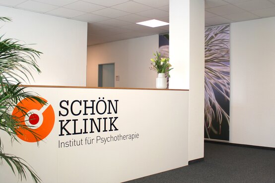 Heller Empfangsbereich mit weißer Theke, Logo „Schön Klinik Institut für Psychotherapie“, Blumen und Pflanzen